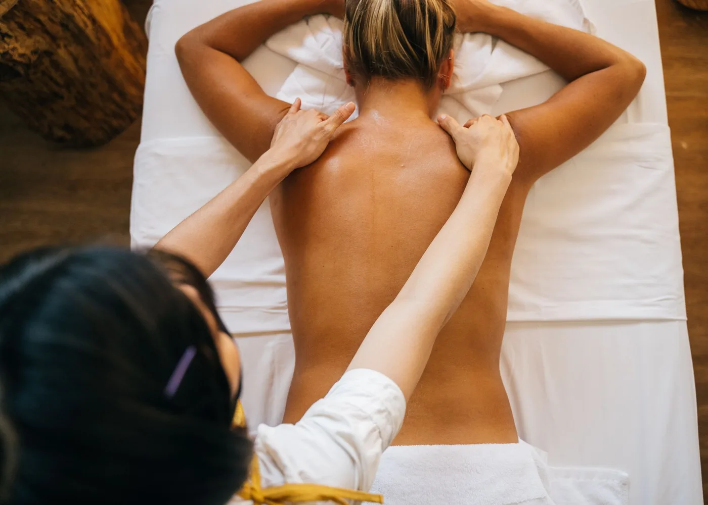 A female therapist pressing gently on the shoulders of a woman lying face down on white linen