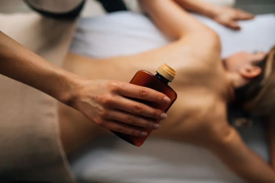 A therapist's hand holding an amber bottle of massage oil above a client lying on her back under a sheet